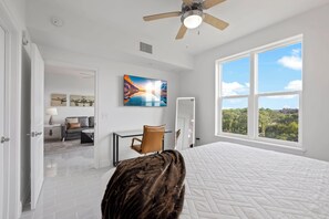 A bedroom with a large bed, a ceiling fan, a desk with a chair, a painting, and a view of the outdoors.