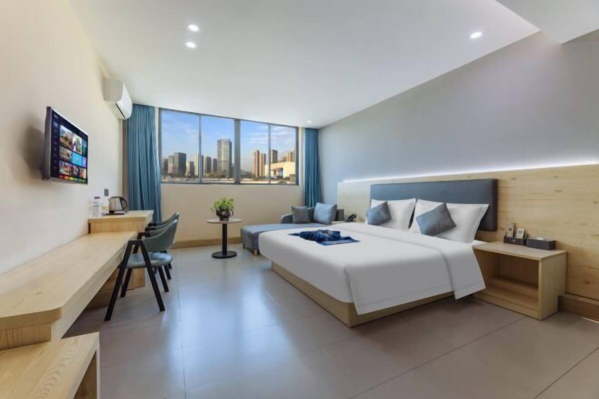 A modern hotel room with a large bed, a desk, a chair, a small table with a plant, and a city view.