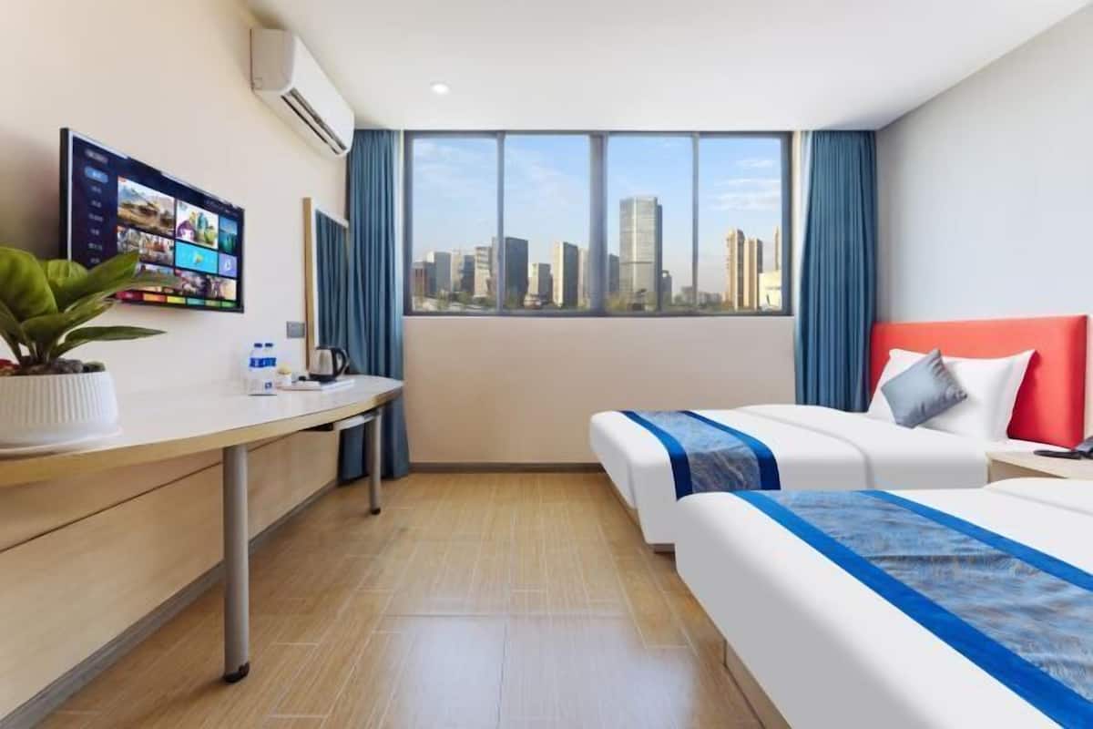 A hotel room with a bed, a desk, a potted plant, a flat-screen TV, and a city view.