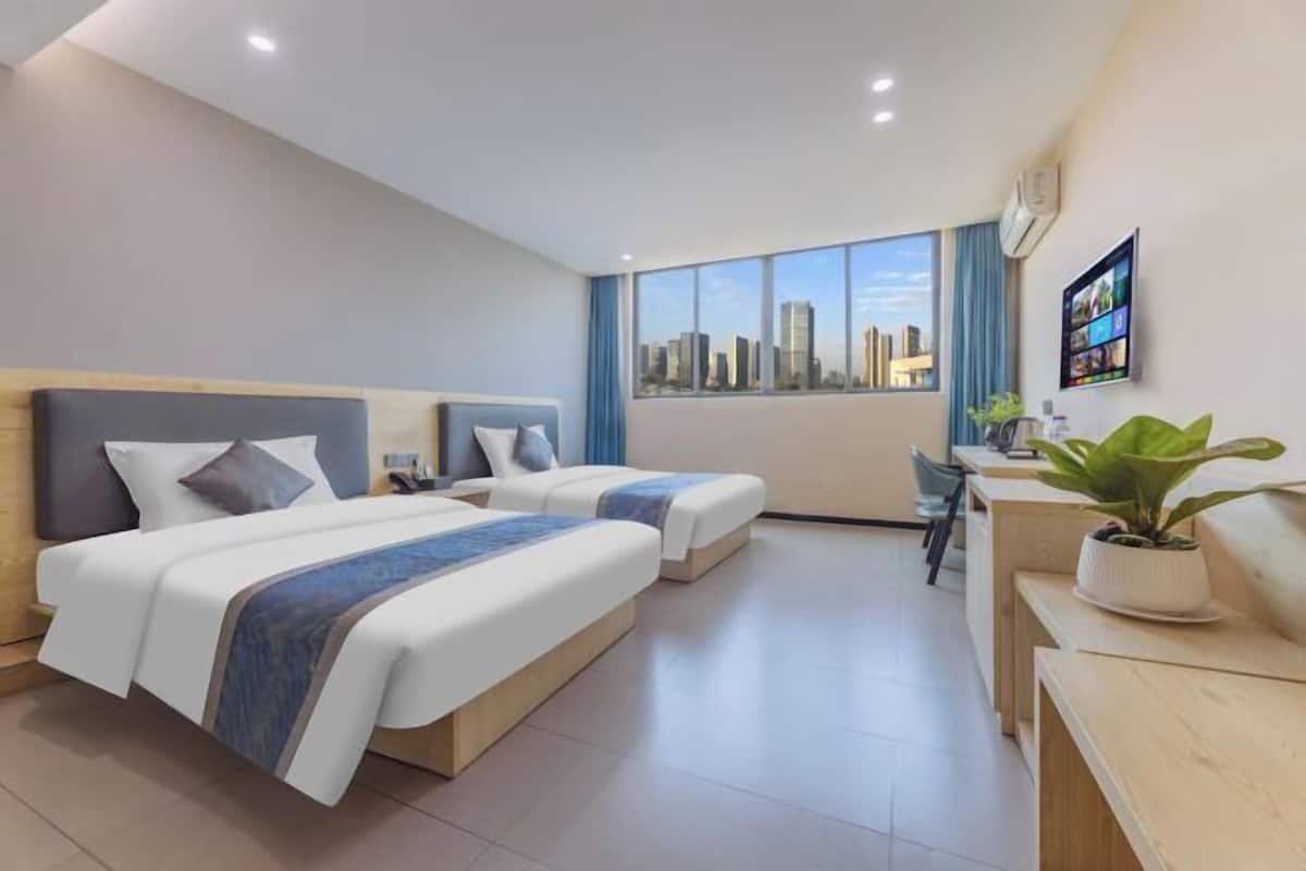 A hotel room with two beds, a desk, a chair, a potted plant, and a city view.