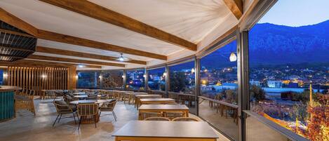 Ein Restaurant mit Holztischen und -stühlen, große Fenster mit Blick auf die Stadt und die Berge im Abenddämmerung.