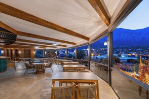 A restaurant with wooden tables and chairs, large windows offering a view of the city and mountains at dusk.