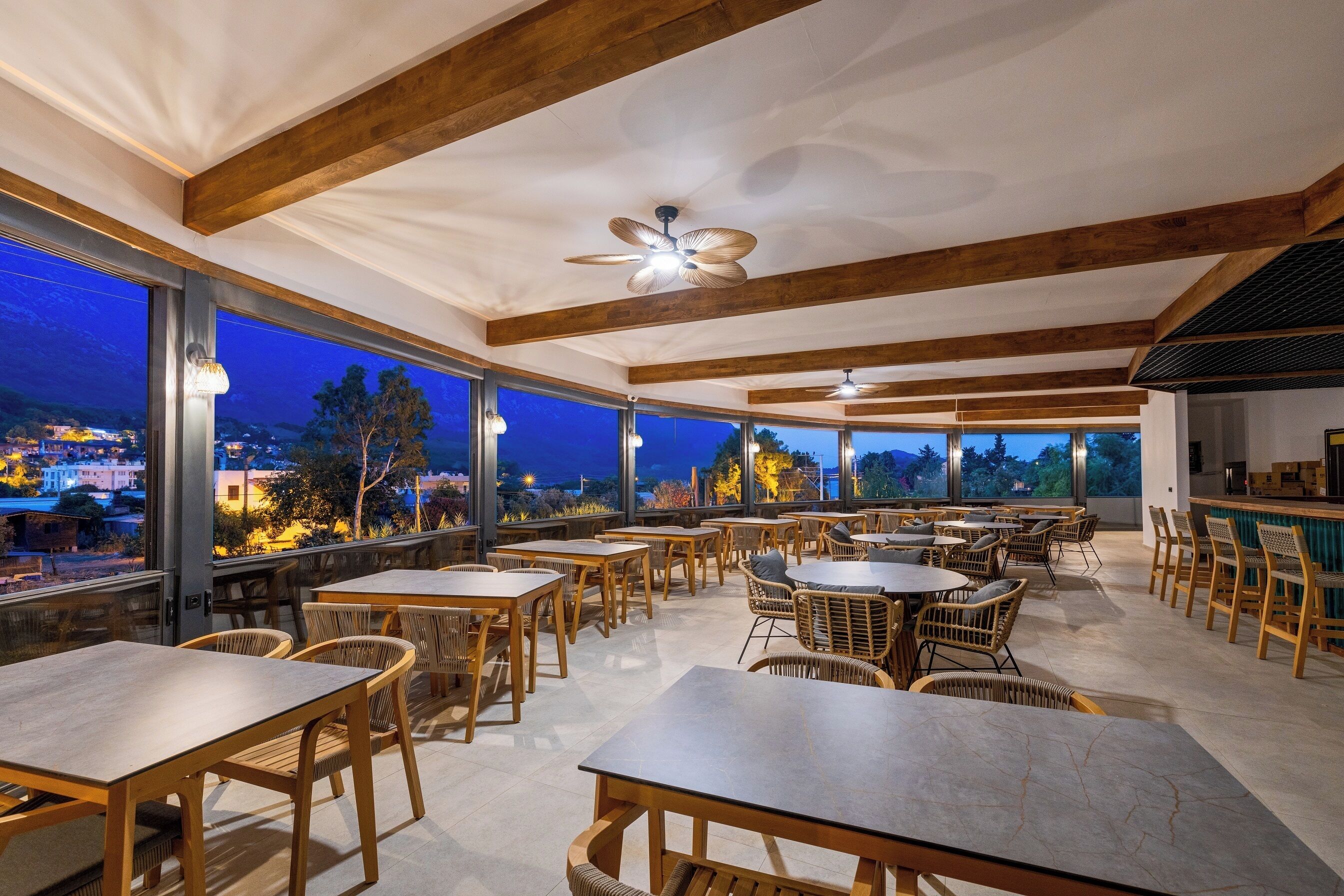Un espace extérieur aménagé pour les repas, avec des tables et des chaises en bois, de grandes fenêtres offrant une vue sur la ville au crépuscule et un ventilateur de plafond.