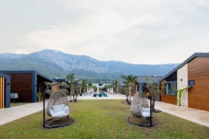 A modern resort with two large egg chairs facing a pool, surrounded by wooden buildings and a mountain view.