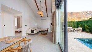 Una sala moderna con sofá, mesa de comedor y sillas, una pequeña mesa redonda, y vistas a una piscina y montañas.