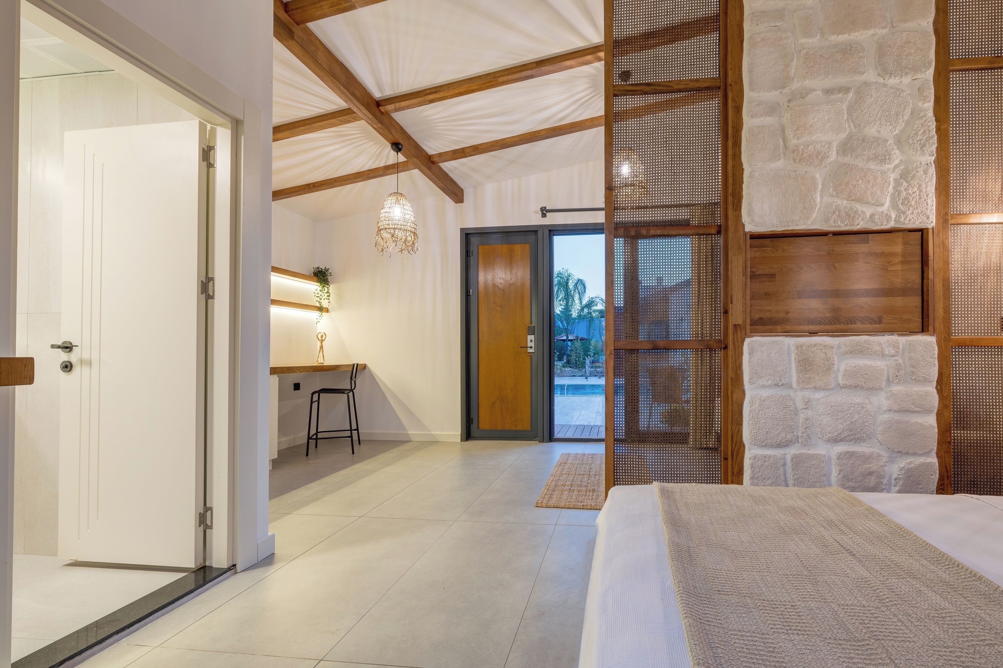 A modern bedroom with a large bed, a wooden ceiling, a stone wall, and a view through a glass door.