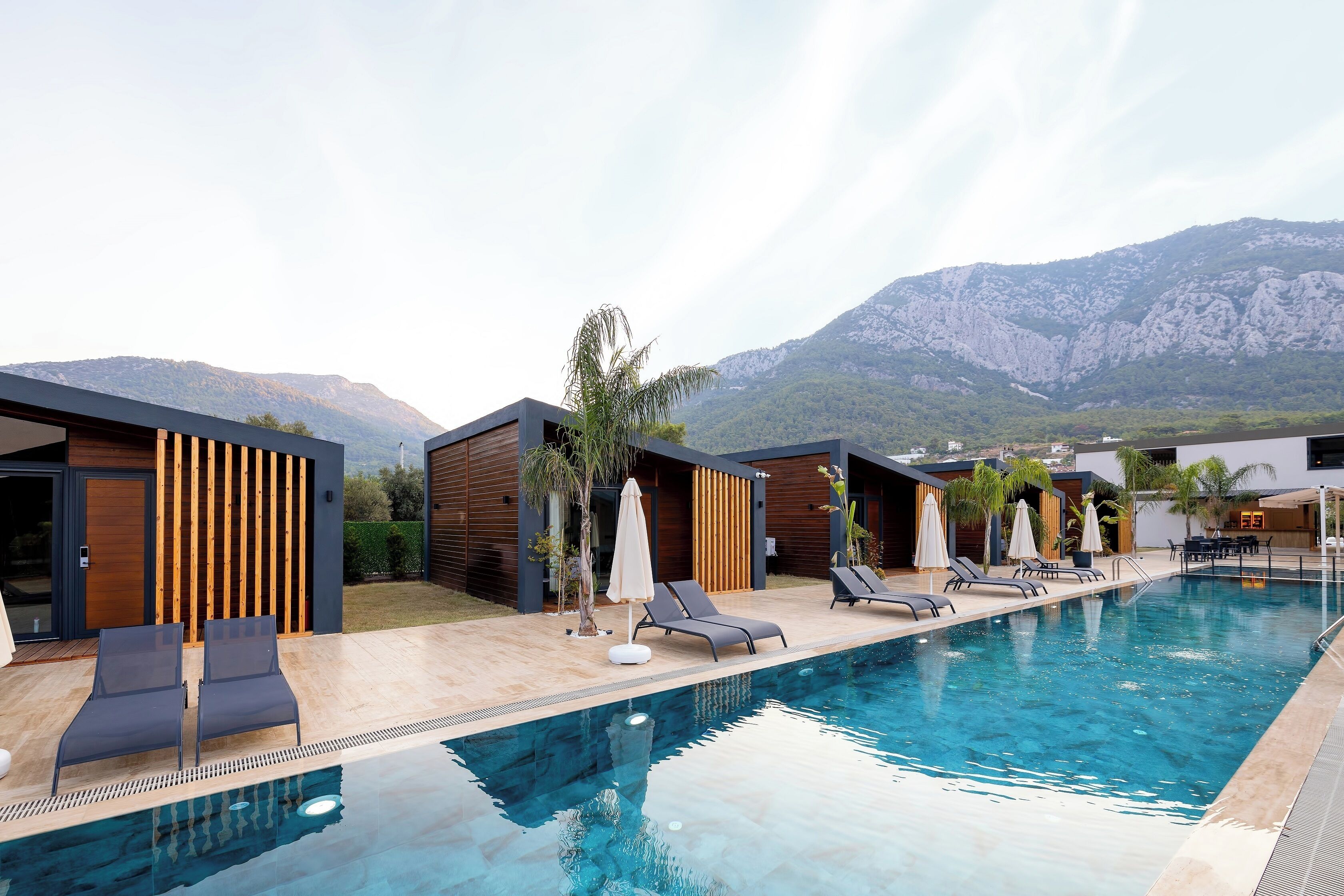 A modern pool area with lounge chairs and umbrellas, surrounded by wooden structures and a mountainous backdrop.