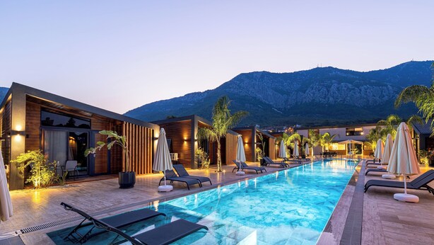 Un área de piscina moderna con sillones de descanso y sombrillas, edificios de madera y montañas al fondo.