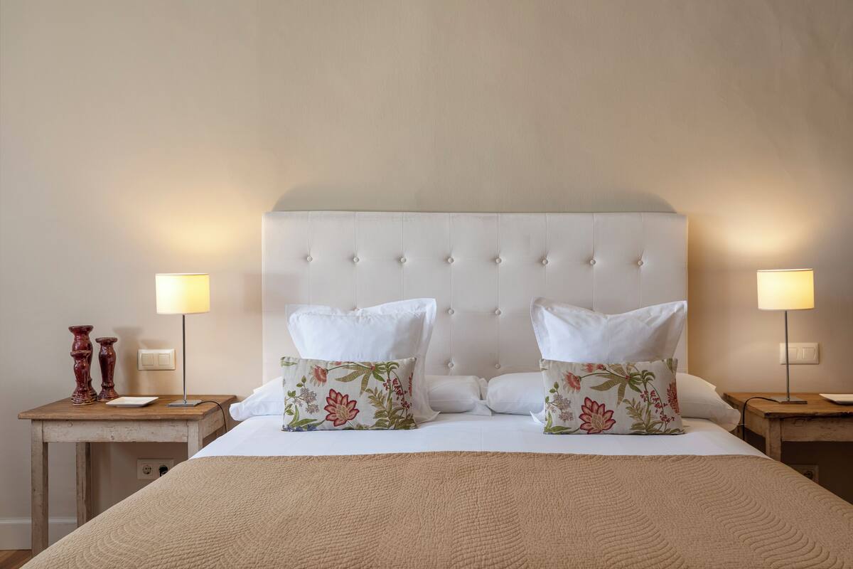 Un lit bien fait, doté d’une tête de lit capitonnée, de deux tables de chevet avec des lampes et d’oreillers à motifs floraux.