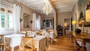 Un espace repas avec des tables et des chaises en bois, un lustre et une vue sur l’extérieur grâce à de grandes fenêtres.