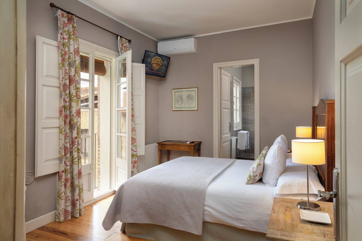 Une chambre à coucher avec un lit, une table de chevet avec une lampe, un téléviseur fixé au mur et une fenêtre avec des rideaux.