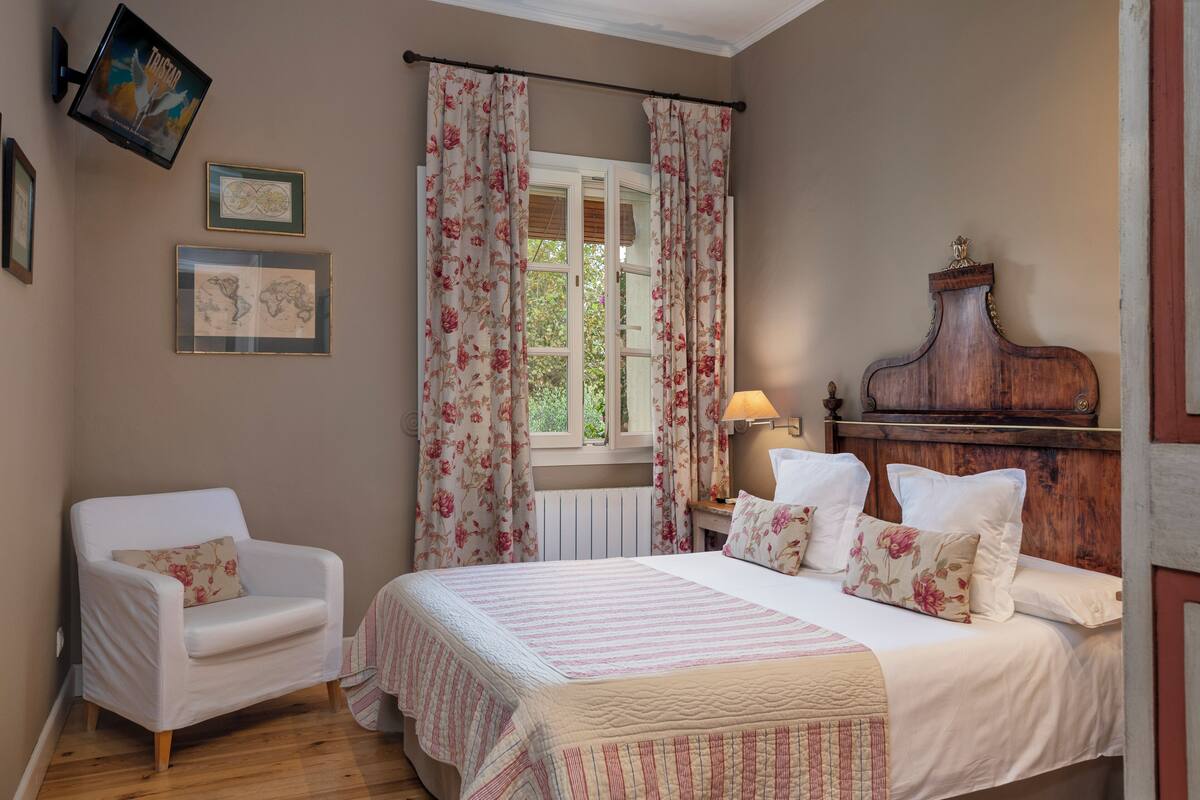 Une chambre à coucher avec un lit, une chaise, une commode et une fenêtre avec des rideaux.