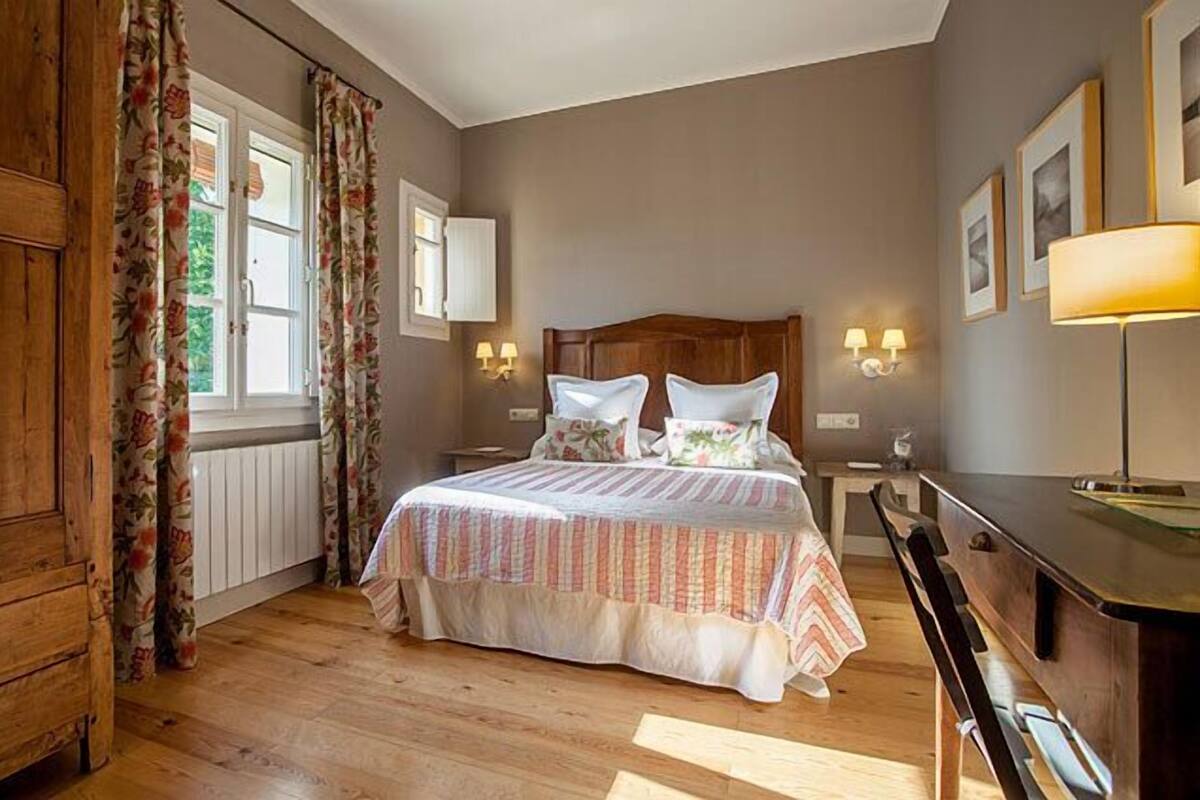 Une chambre à coucher avec une tête de lit en bois, un lit avec une literie à rayures, deux lampes de chevet, une commode et une fenêtre ornée de rideaux à motifs floraux.