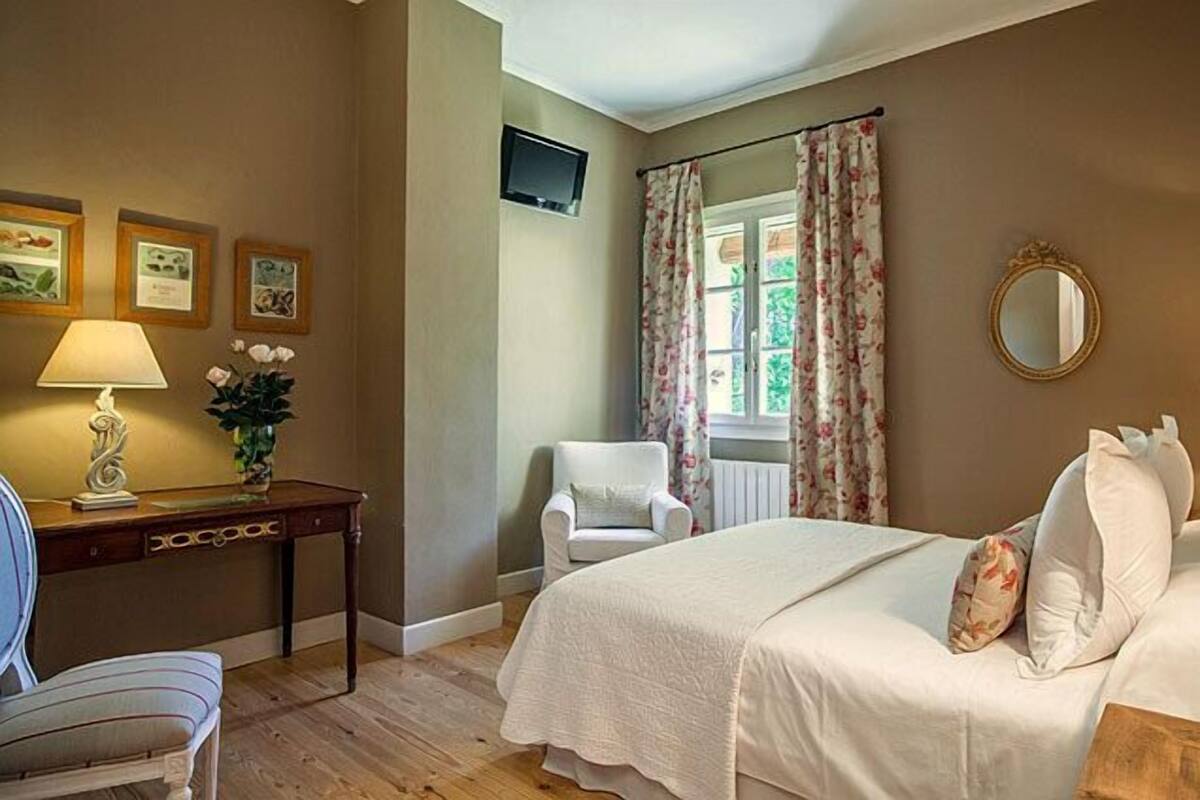 Une chambre à coucher comprenant un lit, une chaise, un bureau, une lampe, un miroir et une fenêtre avec des rideaux.