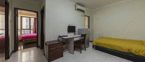 A small, clean room with a bed, a desk, and a chair. There is a window with curtains, an air conditioning unit, and a door leading to another room.