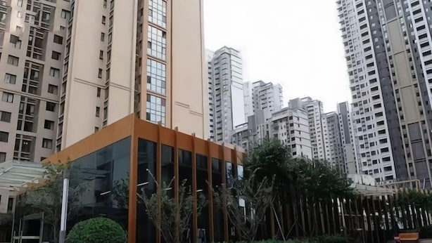 Un edificio moderno con fachada de vidrio, rodeado de vegetación y edificios residenciales altos adyacentes.