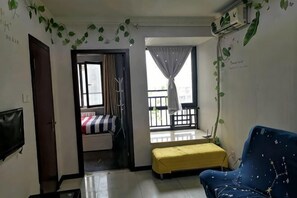 A room with a bed, a yellow bench, a blue chair, a TV, and a balcony with curtains. - Zhengzhou Ruxinjia Apartment (Zhenghong Digital Apartment) (Zhengzhou)