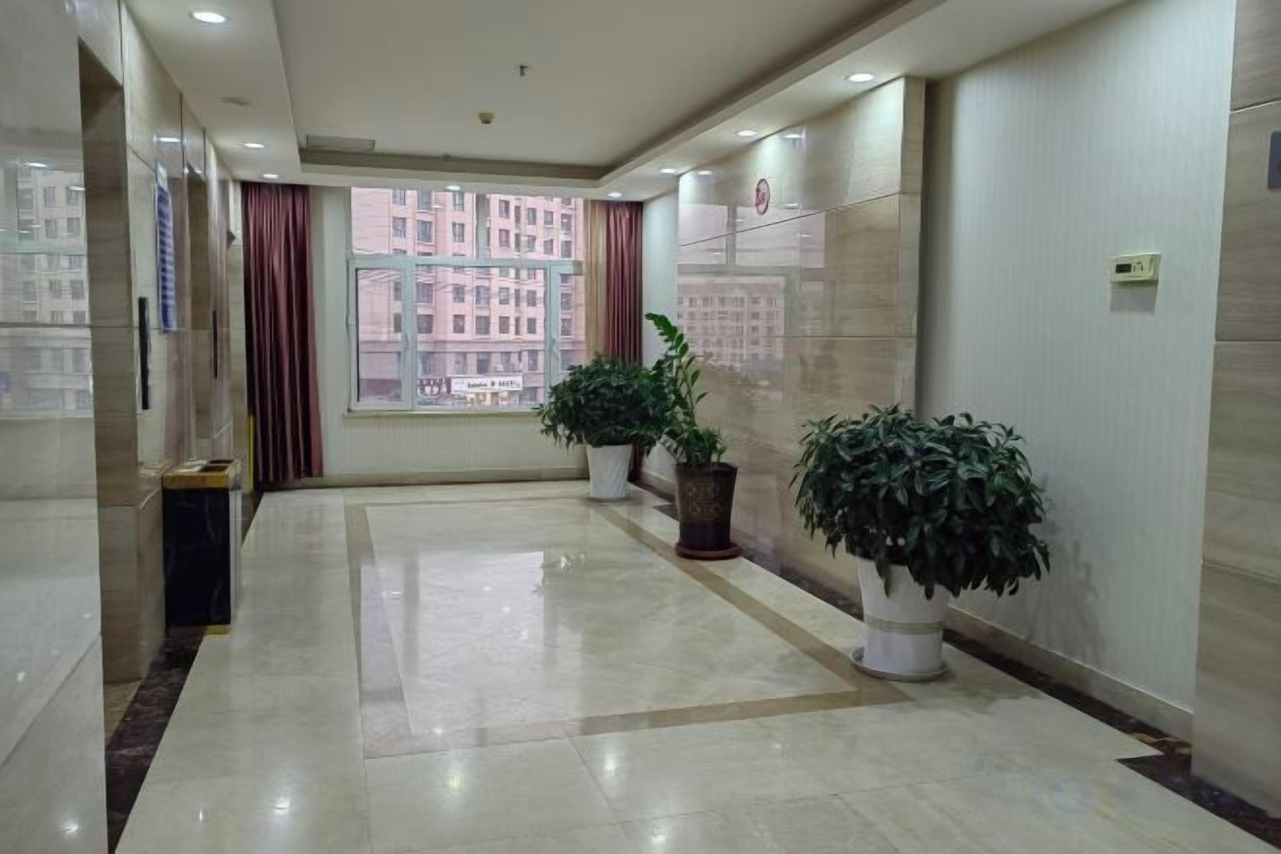 A hotel corridor with a view of buildings outside, potted plants, and a tiled floor.