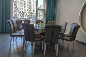 A dining area with a round glass table and six chairs, large windows with curtains, and a view of buildings outside. - Times Lijing Hotel Xilinhot (Xilinhot)