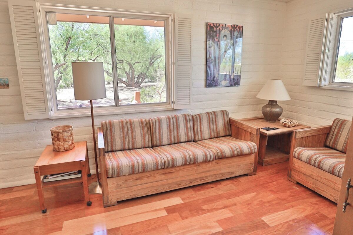A cozy living room with a sofa, armchairs, a coffee table, and a lamp. There is a window with blinds, a painting on the wall, and a small wooden table with a lamp.
