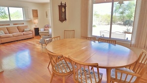 A spacious living room with a round wooden table and chairs, a sofa, a clock on the wall, and large windows offering a view of the outdoors. - Happy Trails Hideaway (Tucson)