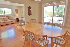 A spacious living room with a round wooden table and chairs, a sofa, a clock on the wall, and large windows offering a view of the outdoors. - Happy Trails Hideaway (Tucson)