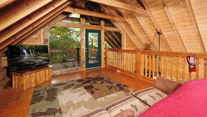 An attic bedroom with a wooden ceiling, a television on a wooden stand, a patterned rug, and a view of the outdoors through a door. - 3 BR/3 bath (sleeps 8) located between GBurg & PF! Hot tub, pool table, privacy! (Sevierville)