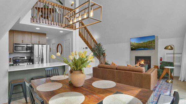 Een moderne keuken met roestvrijstalen apparatuur, een eethoek met een houten tafel en krukken, een woonkamer met een open haard, en een trap met houten leuningen.
