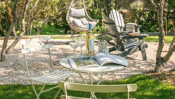 A round table with a magazine, two wine glasses, and a bottle of wine in a garden setting with outdoor furniture. - Sardegna è - Beach Rooms (Olbia)