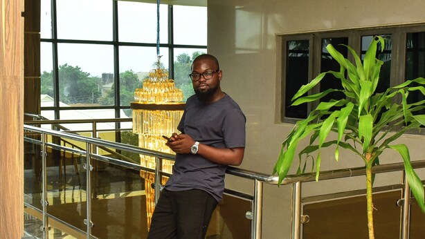 A man standing by a railing in a lobby with a large potted plant, a chandelier, and a view of the outdoors through large windows.