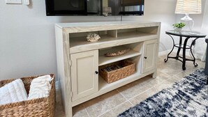 A living room with a flat-screen TV mounted on the wall, a light-colored wooden cabinet with open shelves, a small round table with a lamp, and a blue and white patterned rug on the floor.