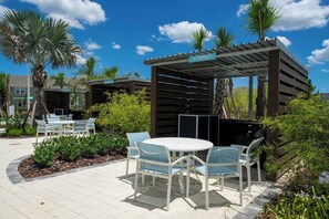 An outdoor patio area with a round table and chairs, surrounded by greenery and palm trees. - Stunning 7Bd Pool Jacuzzi near Disney Resort 16217 (Clermont)