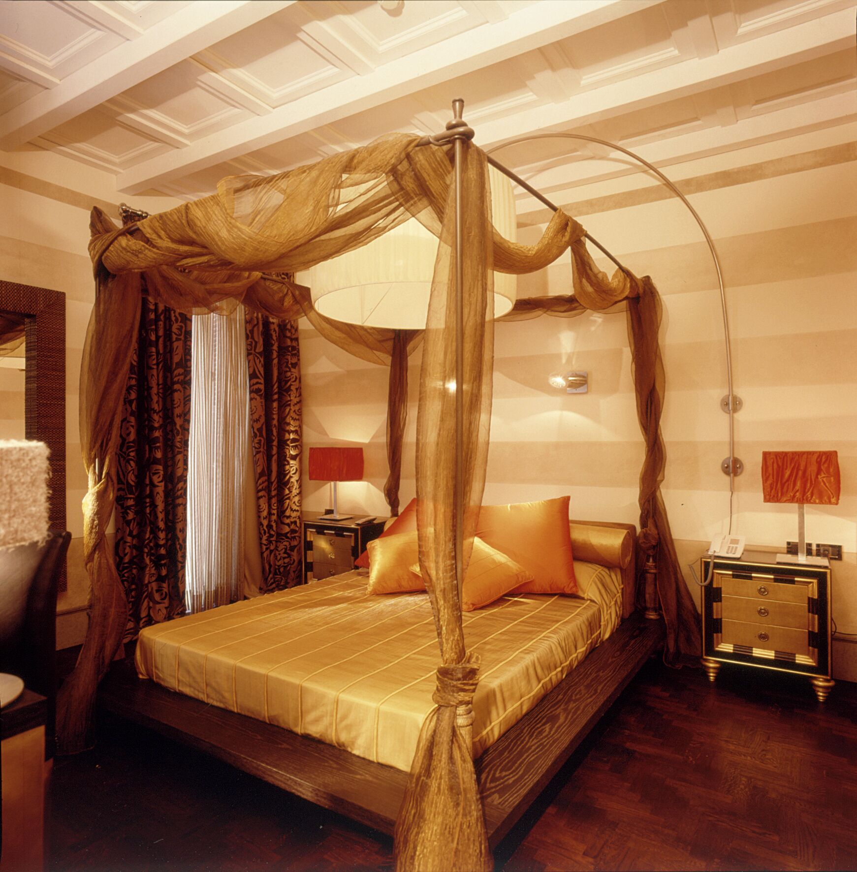 A four-poster canopy bed with a wooden frame and gold bedding, a nightstand with a telephone, and a lamp on the side table.