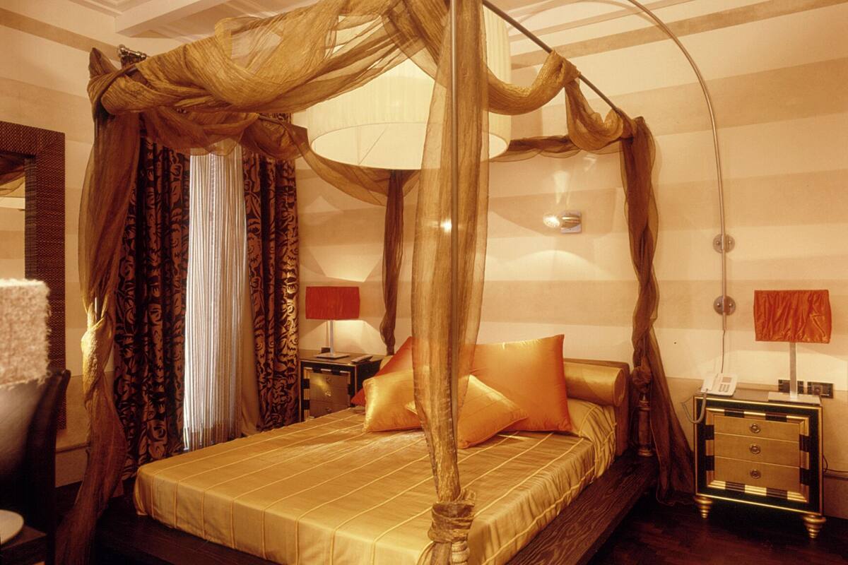 Un lit à baldaquin à quatre poteaux, avec une structure en bois et une literie dorée, une table de chevet avec un téléphone et une lampe sur la table d’appoint.