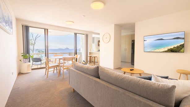 A modern living room with a large sofa, a dining area, and a kitchen. There is a large window offering a view of the sea. - The Point 13, 5-7 Mitchell St - stunning water views, complex pool and WiFi (Soldiers Point)