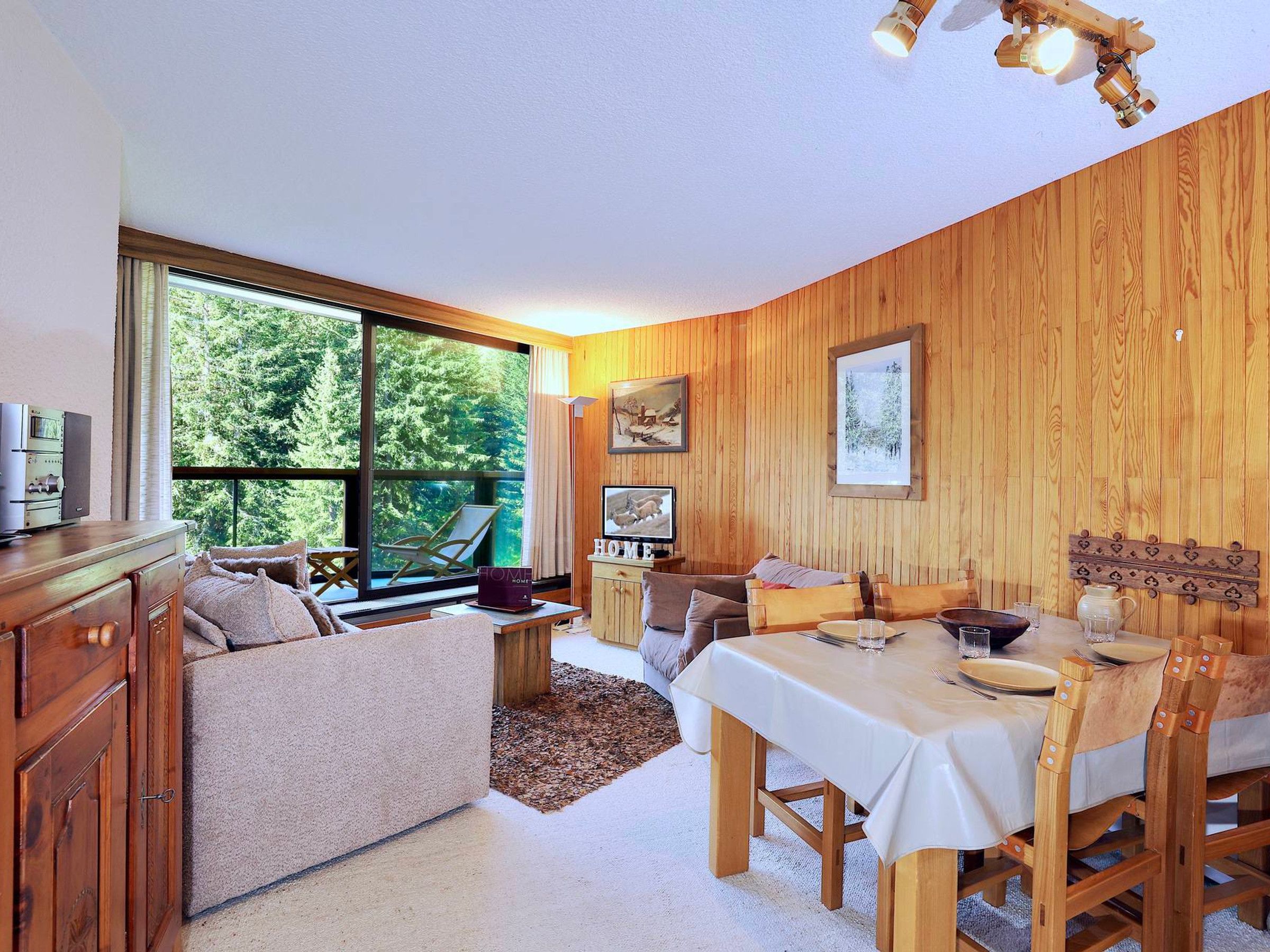 A cozy room with a large window, wooden walls, a dining table with chairs, a sofa, and a cabinet.