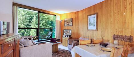A cozy room with a large window, wooden walls, a dining table with chairs, a sofa, and a cabinet.