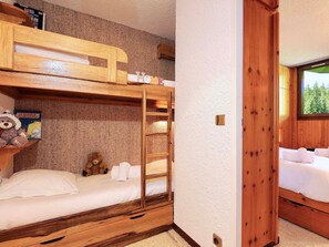 A bunk bed room with a wooden ladder, a shelf with books and a teddy bear, and a window with a forest view.
