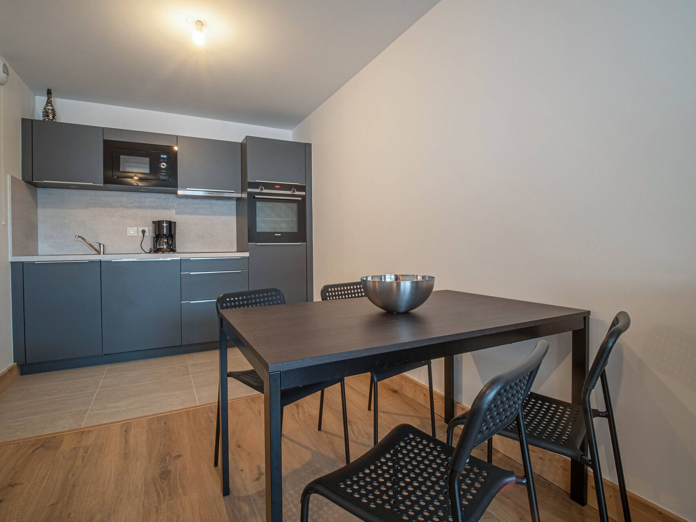 A modern kitchen with grey cabinets, a stainless steel sink, and a coffee machine. There is a wooden dining table with four chairs.