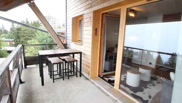 A wooden balcony with a table and stools, a sliding glass door, and a view of trees and buildings.
