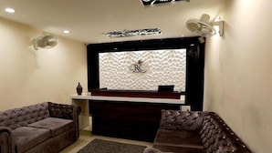 A room with a dark brown sofa, a smaller brown sofa, and a dark brown ottoman. There is a white textured wall with a logo and a small white table with a vase. - Hotel RK Grande (Ludhiana)