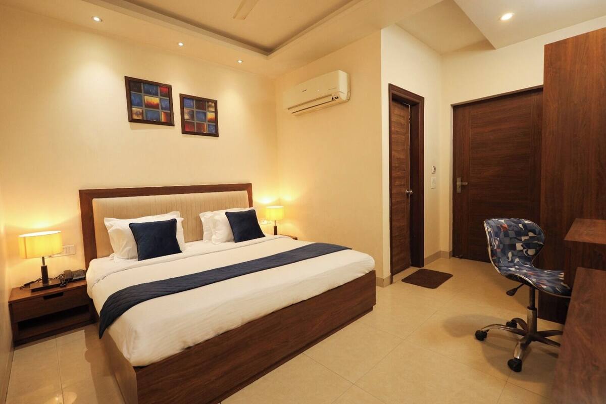 A hotel room with a large bed, bedside lamps, an air conditioning unit, a desk with a chair, and a door.