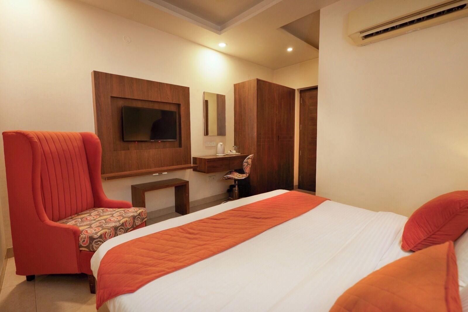 Habitación de hotel con una cama, un sillón rojo, un escritorio pequeño, un espejo y un televisor fijado a la pared.