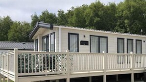 A single-story mobile home with a balcony, a paved driveway, and a surrounding grassy area. - The Westwood at Percy Wood (Morpeth)