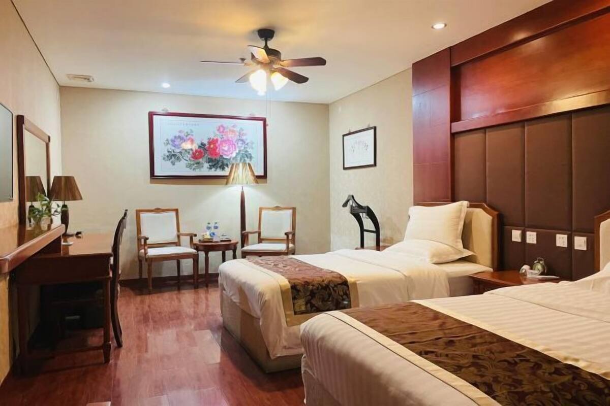 Une chambre d’hôtel avec deux lits, un bureau, des chaises, un ventilateur de plafond et des œuvres d’art accrochées aux murs.
