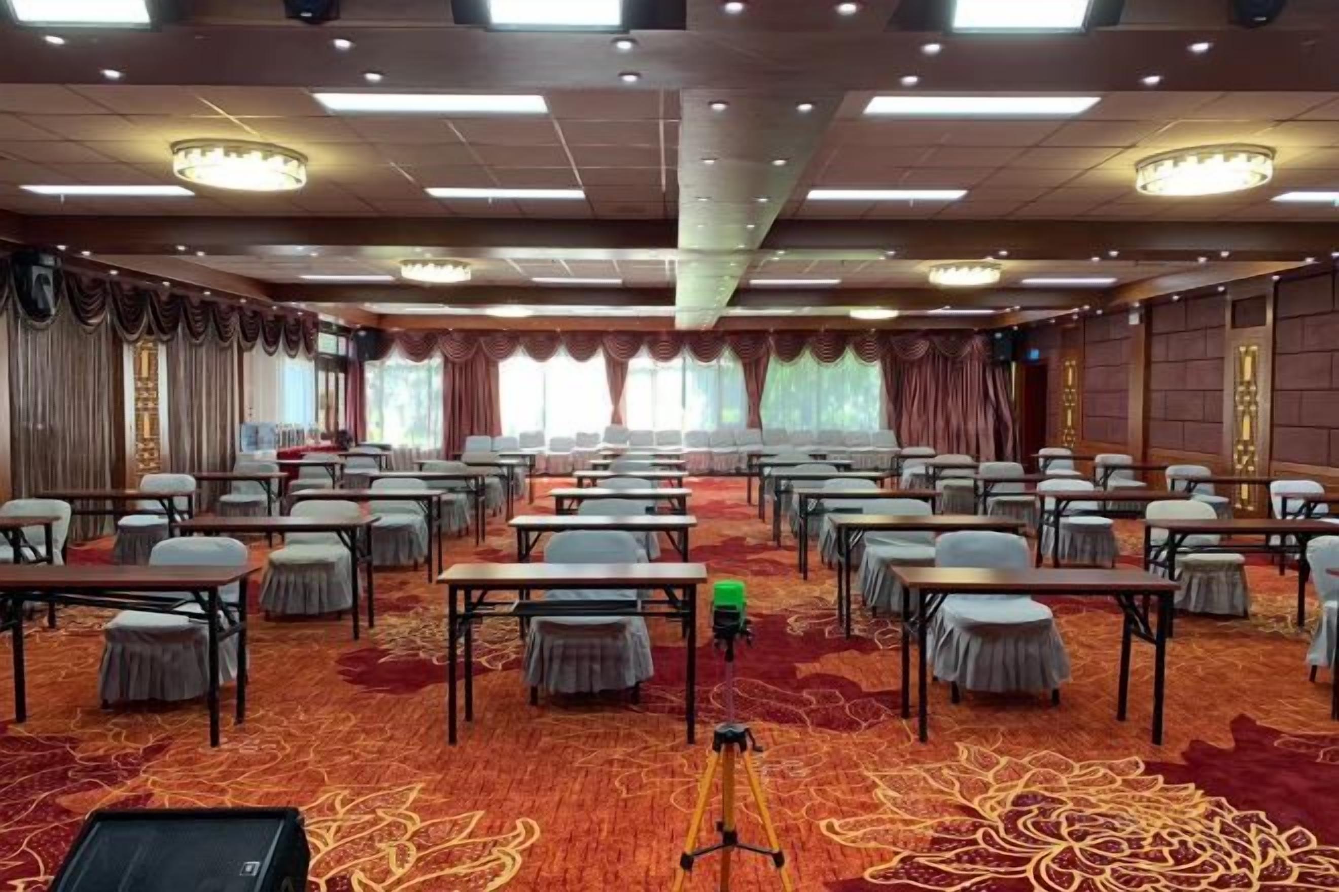 Un salón de conferencias con filas de mesas y sillas, piso alfombrado y un ventanal con cortinas.