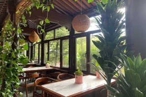 Een eetruimte binnenshuis met houten tafels en stoelen, grote ramen en hangende planten.