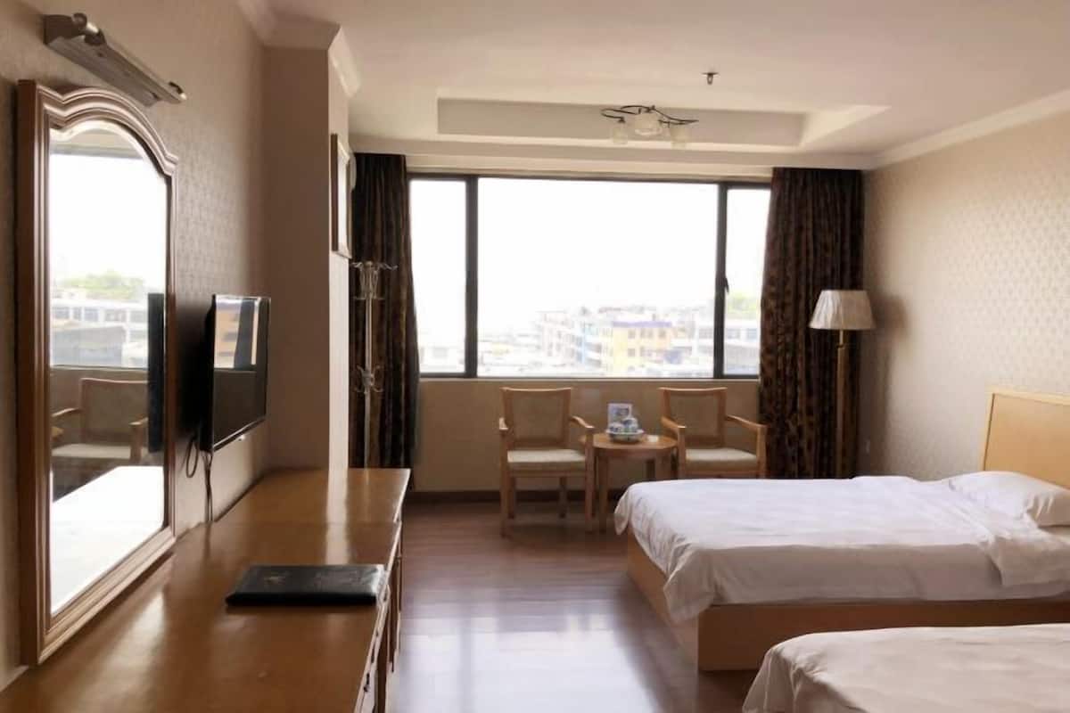 Ein Hotelzimmer mit zwei Betten, einem Schreibtisch, einem Stuhl, einem Fernseher und einem großen Fenster mit Vorhängen.