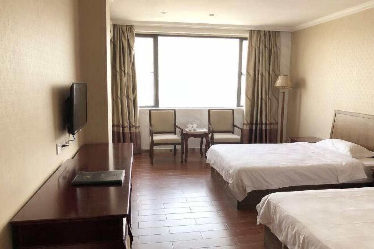 Ein Hotelzimmer mit zwei Betten, einem Schreibtisch, einem Fernseher, einem Fenster mit Vorhängen und einem kleinen Tisch mit Stühlen.