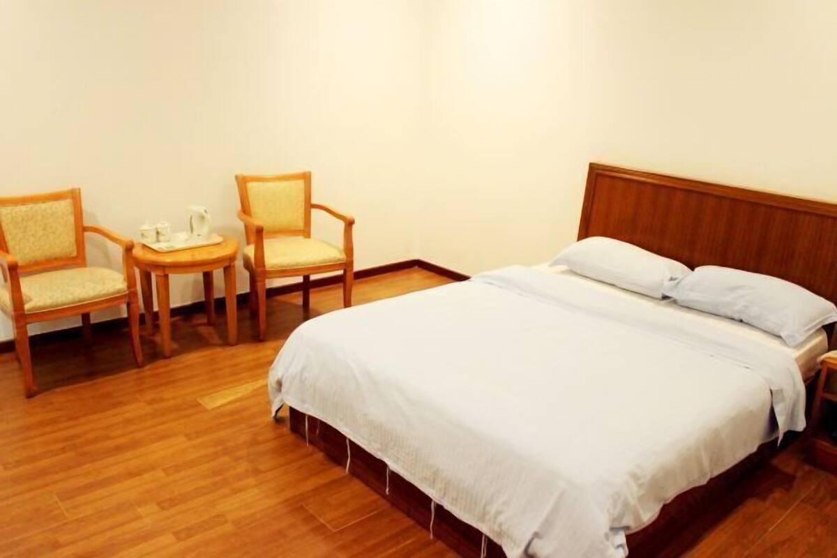 Ein Hotelzimmer mit einem hölzernen Bett, zwei Stühlen, einem kleinen Tisch mit Tablett und einer weißen Wand.